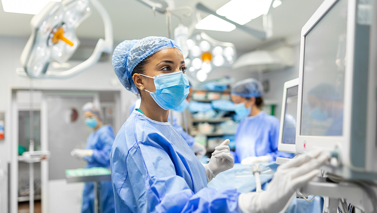 Anesthetist Working In Operating Theatre Wearing Protective Gear checking monitors while sedating patient before surgical procedure in hospital.